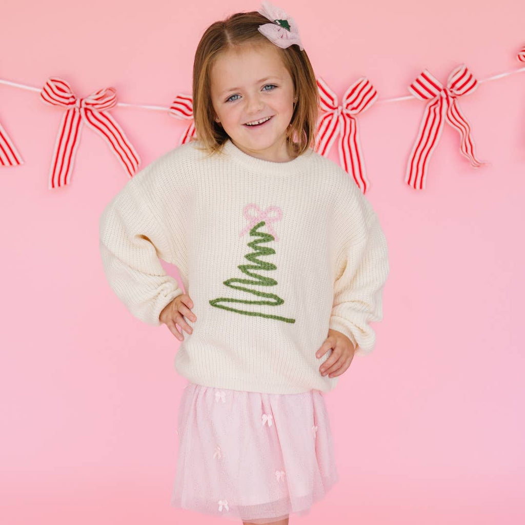 Child wearing a cream sweater with a Christmas tree design on a pink background with decorative bows.