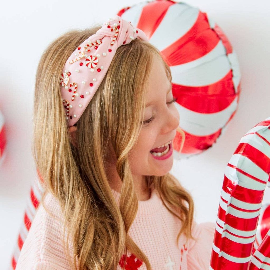 Young girl wearing a pink headband with candy cane decorations, standing in front of candy cane-themed balloons.