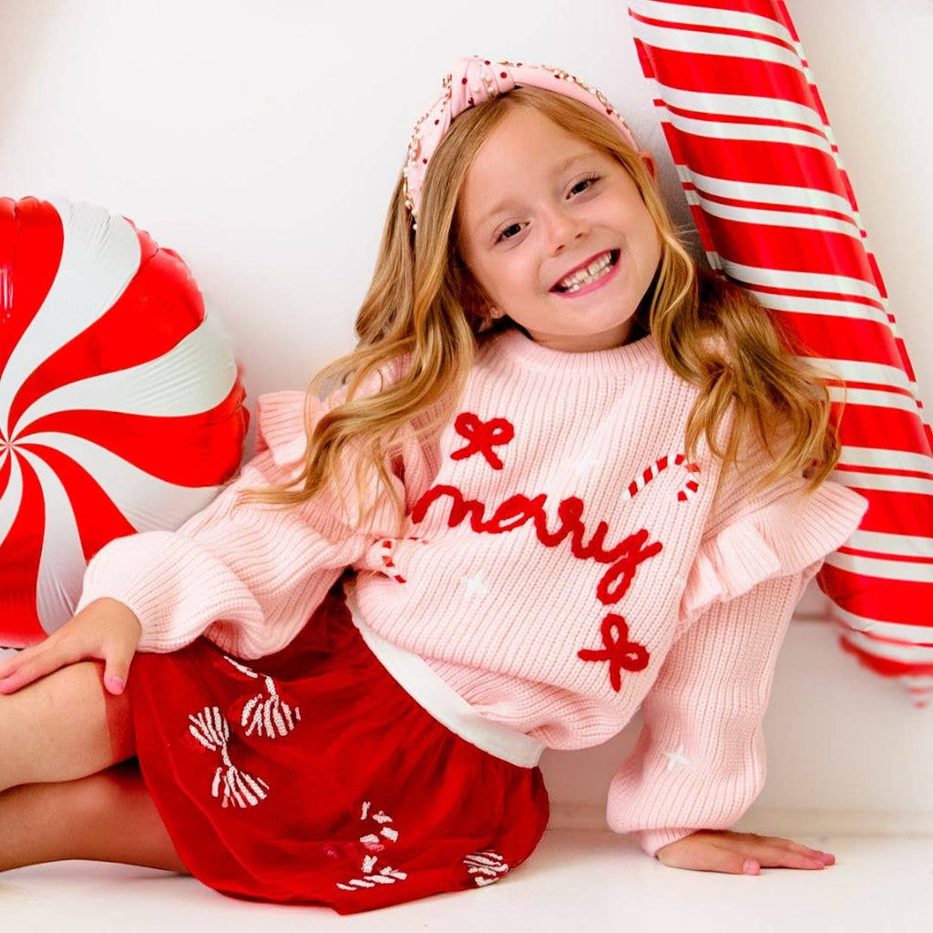 Young girl in a pink sweater with 'Merry' and red skirt sitting next to candy cane-themed decorations.