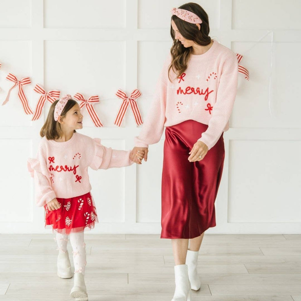 Two people wearing matching pink and red outfits with 'Merry' text, standing against a white wall with decorative ribbons.