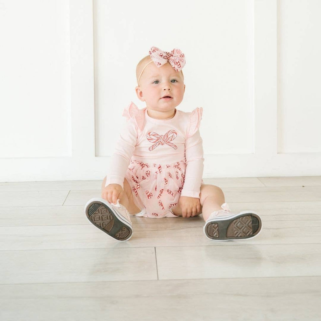 Baby sitting on a light wooden floor wearing a pink outfit with a bow and shoes.