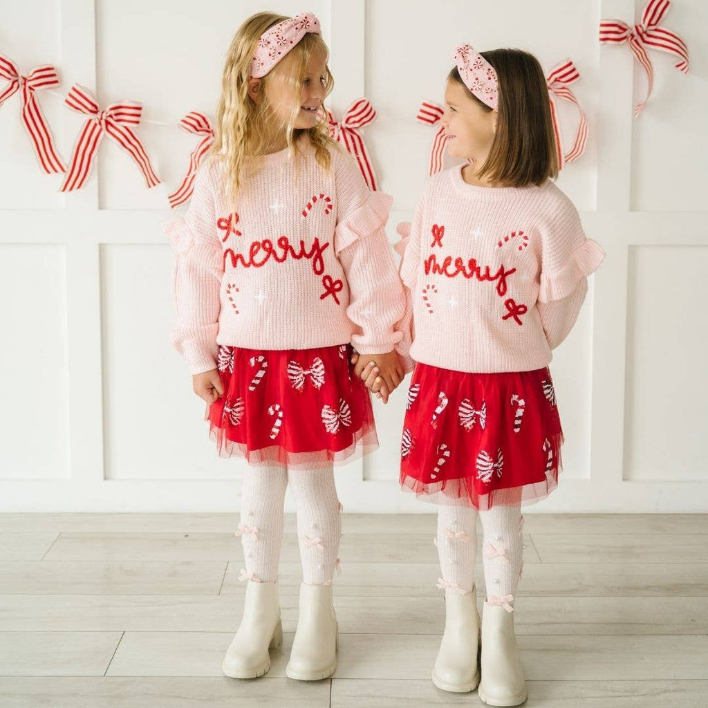 Two children wearing matching pink sweaters with 'Merry' text and red skirts, standing against a white wall with decorative bows.