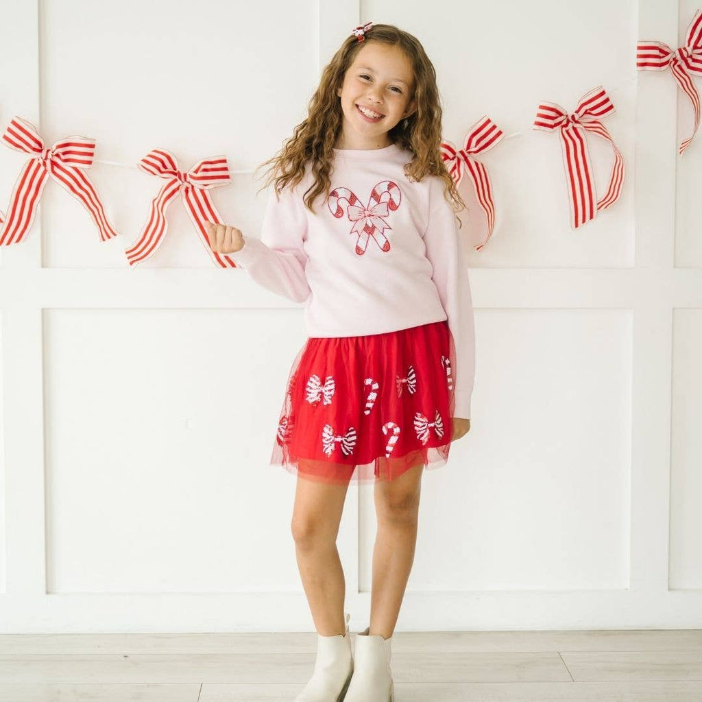 Young girl wearing a white sweater and red skirt with candy cane designs, standing in front of a white wall with red bow decorations.