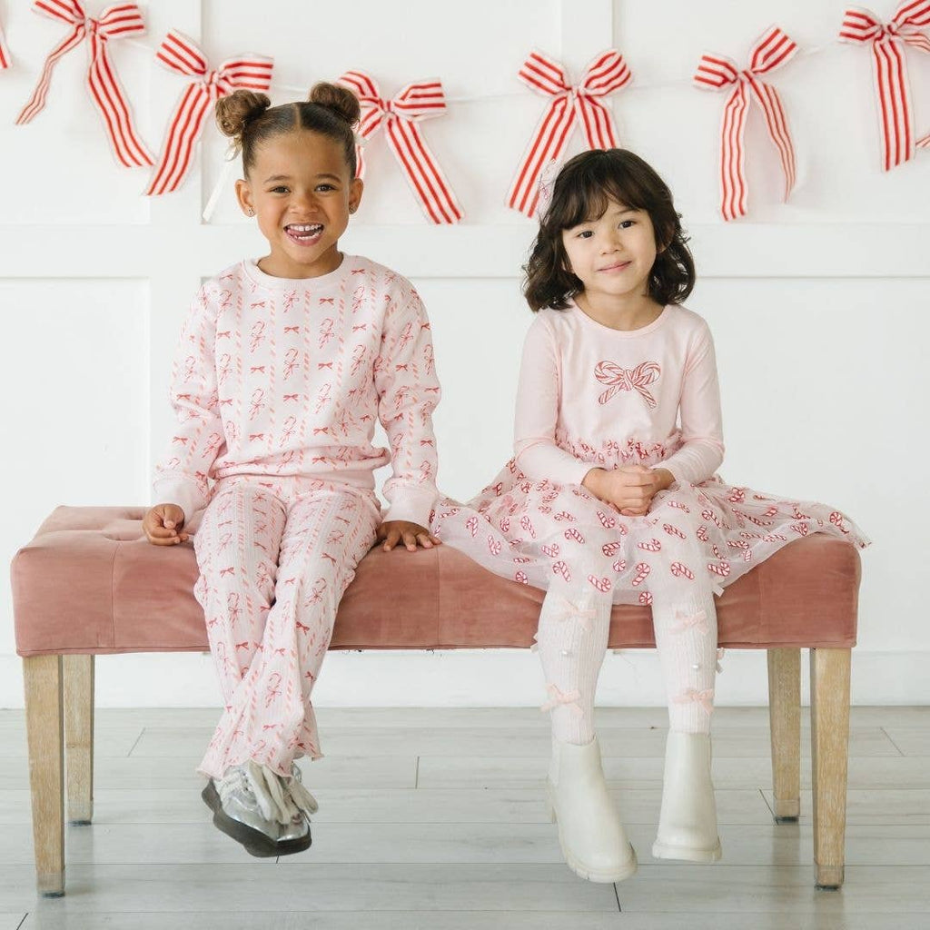 Two young girls in matching pajama sets sitting on a pink bench with decorative bows in the background.