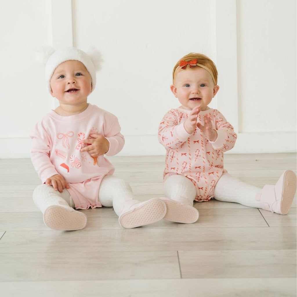 Two babies sitting on a light wooden floor wearing matching pink outfits with white pants.