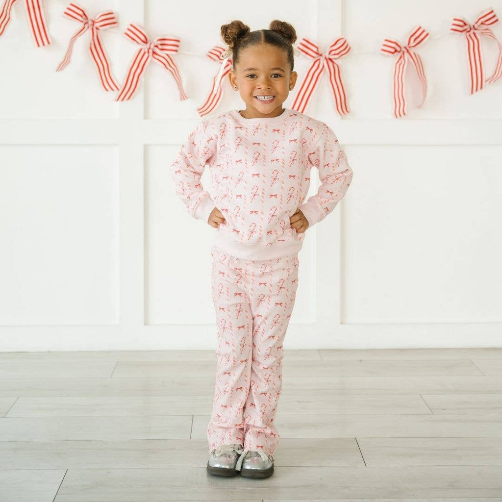 Child wearing pink pajamas with a pattern, standing in front of a white wall with red bow decorations.