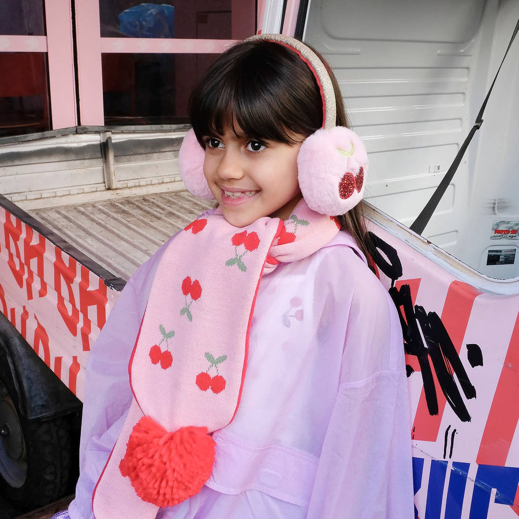 Child wearing pink earmuffs and a cherry-patterned scarf in front of a colorful vehicle.