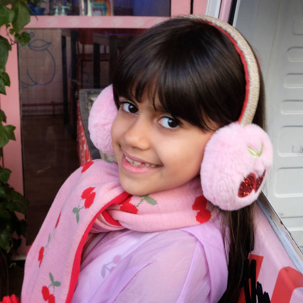 Young girl wearing pink earmuffs and a matching scarf indoors.