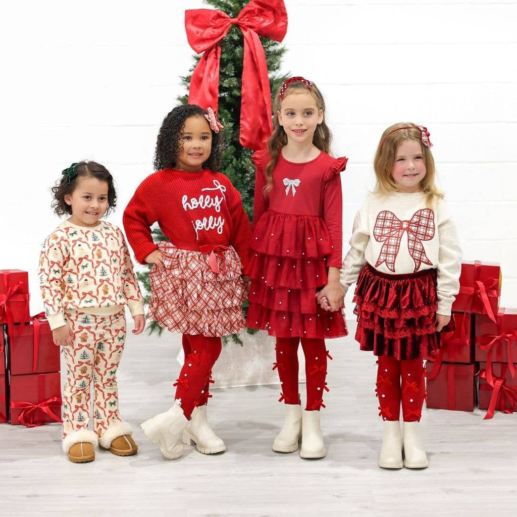 Four children in festive Christmas outfits standing in front of a decorated Christmas tree with red bows.