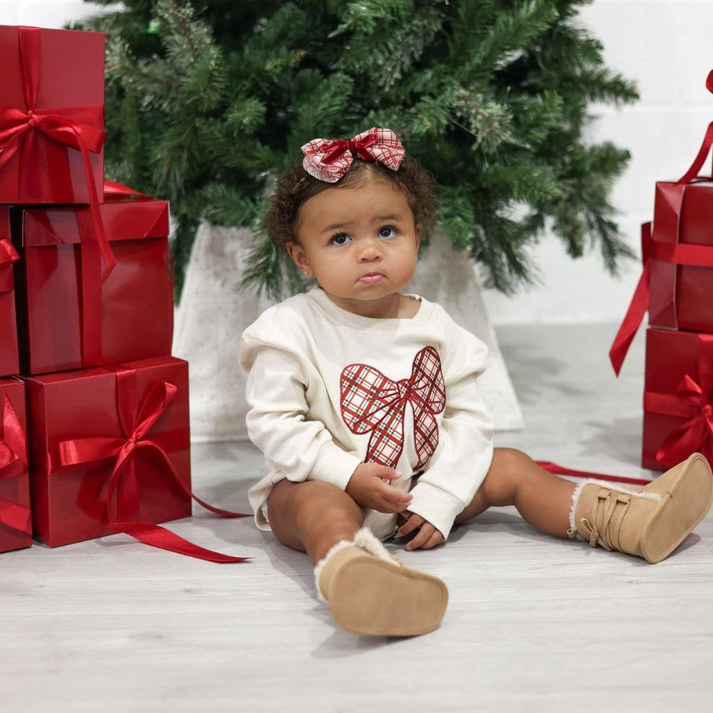 Child in a white outfit with a red bow sitting among Christmas presents and a tree.
