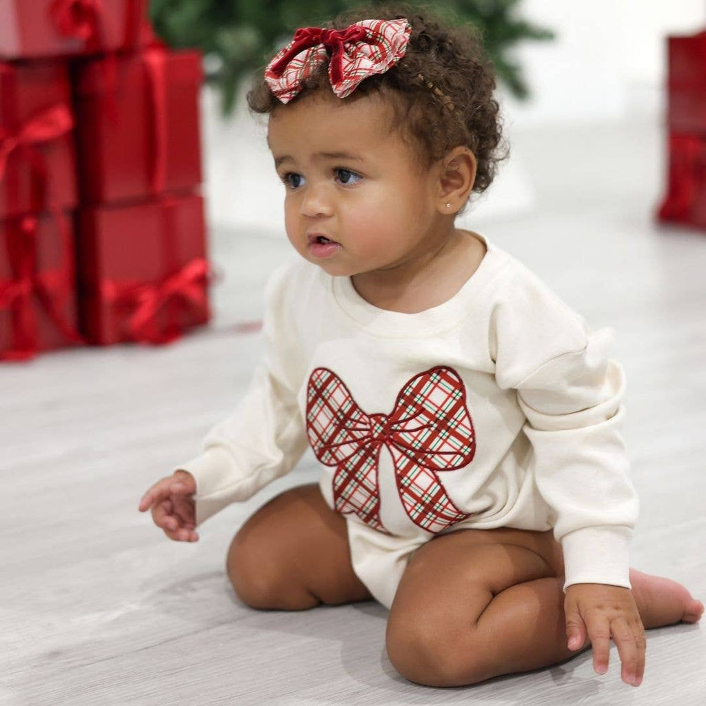 Baby wearing a white onesie with a red bow design, sitting on a light-colored floor with red gift boxes in the background.