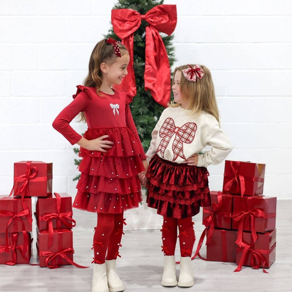 Two children in matching red and white outfits standing in front of a Christmas tree with red bows and presents.