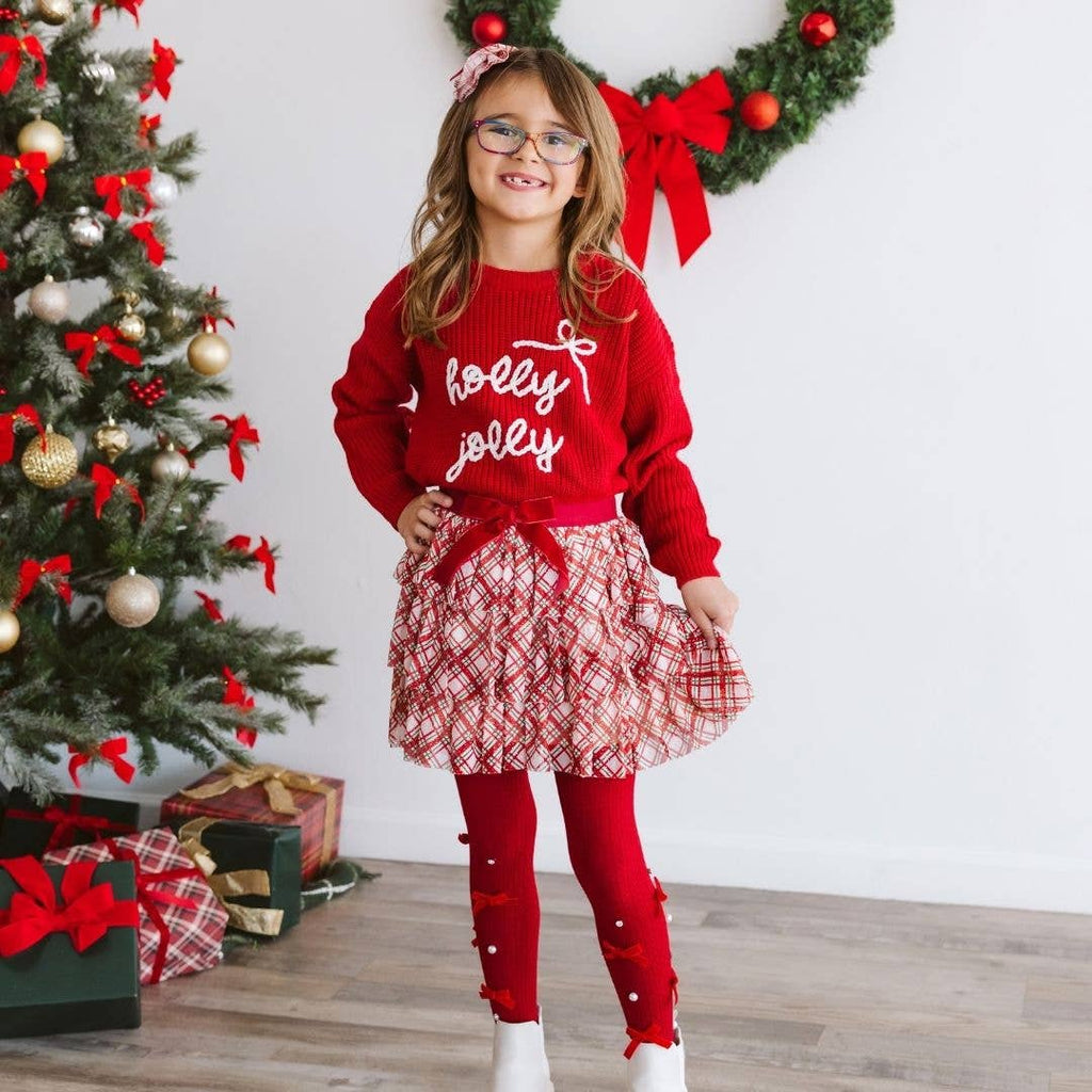 Child wearing a red sweater with 'holly jolly' text, standing in front of a decorated Christmas tree.