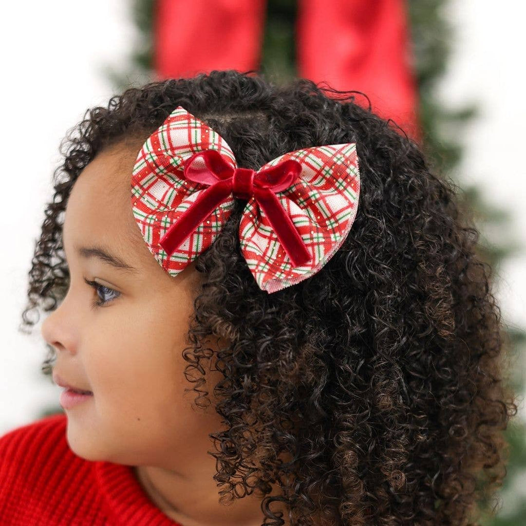 Child with curly hair wearing a red and green plaid bow in front of a blurred Christmas tree.