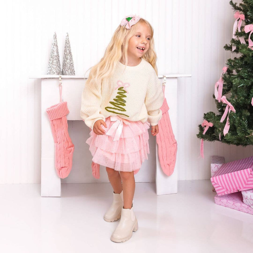 Young girl in a festive setting with Christmas decorations, wearing a white sweater with green text and a pink skirt.