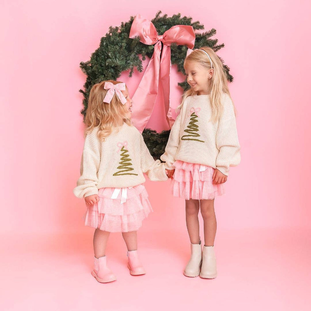 Two children in matching outfits standing in front of a Christmas wreath on a pink background