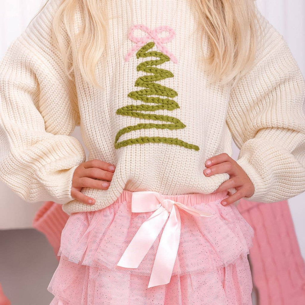 Child wearing a cream sweater with a green knitted tree design and pink skirt.