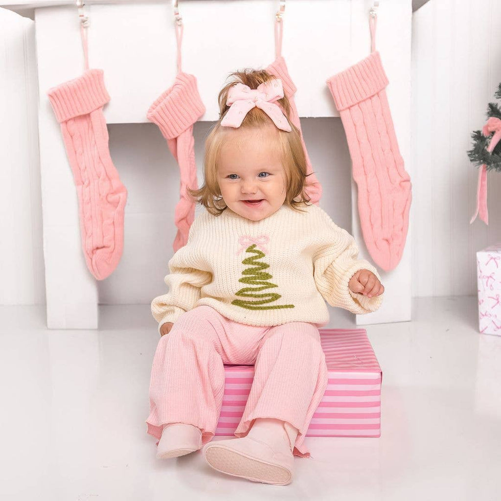Child in a white sweater with a green tree design and pink pants sitting on a striped box, surrounded by pink stockings.