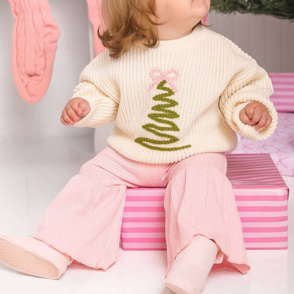 Baby wearing a cream sweater with a green design and pink pants sitting on a pink box.