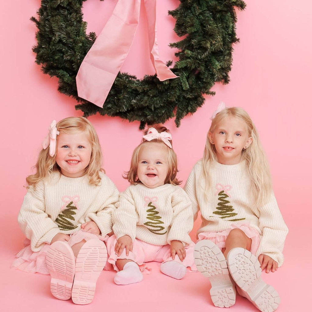 Three children wearing matching Christmas-themed sweaters and skirts sitting in front of a pink background with a wreath.