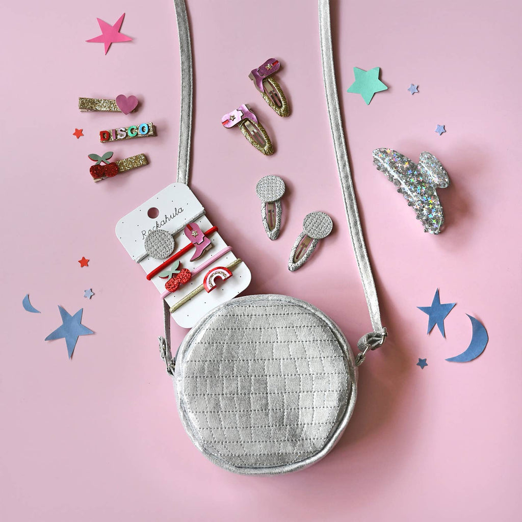Small round silver bag with decorative hair clips on a pink background with star and moon shapes.