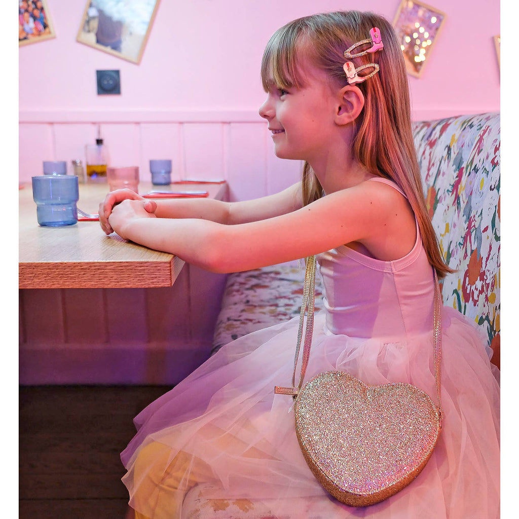 Young girl in a pink dress with a glittery heart-shaped bag sitting at a table.