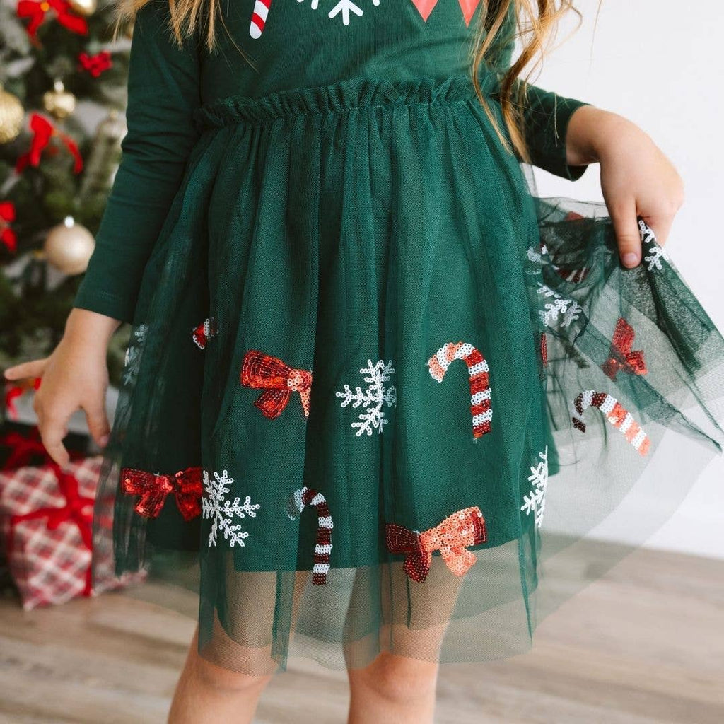 Child wearing a green dress with festive patterns in front of a Christmas tree.