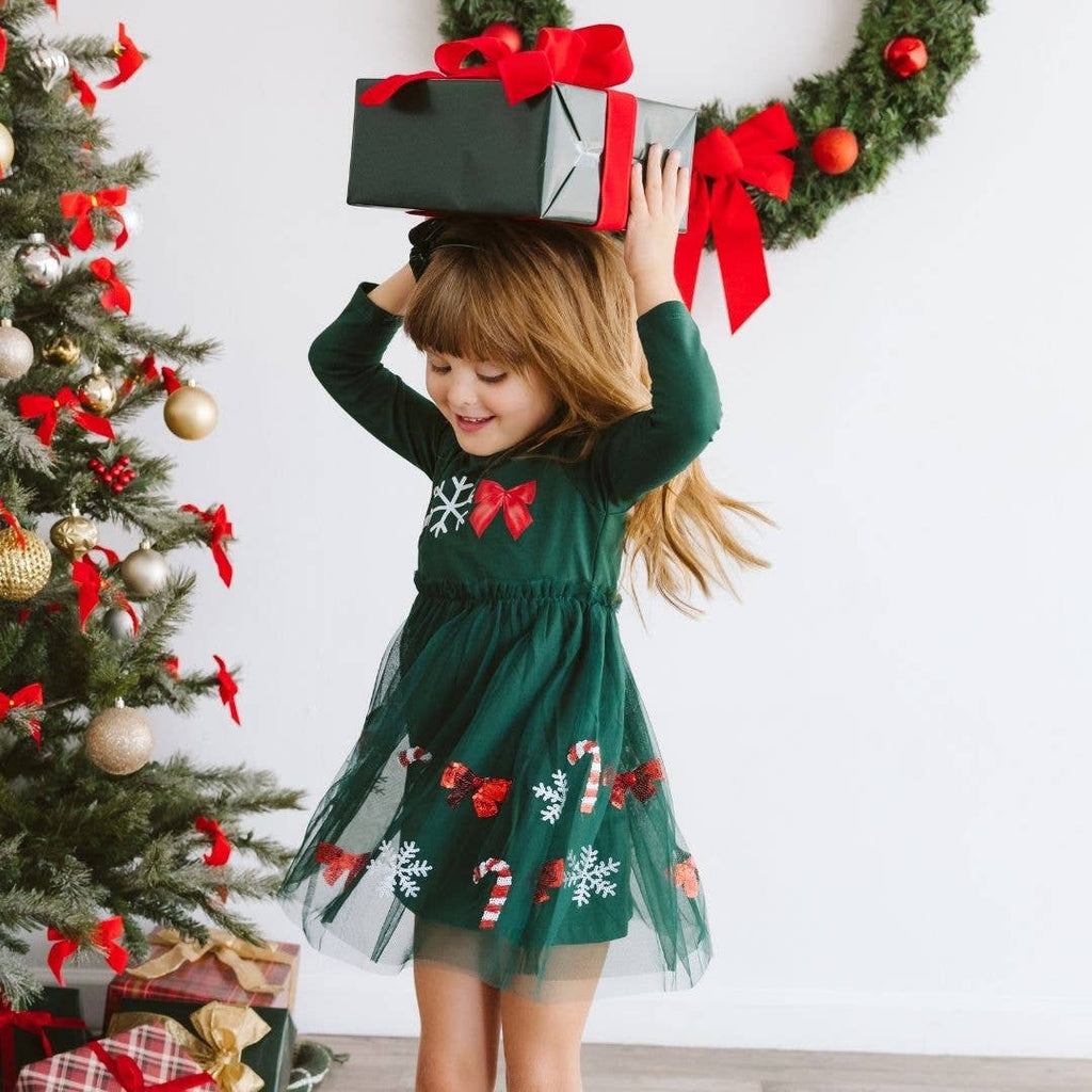 Child in a green Christmas-themed dress holding a gift box with a red ribbon in front of a decorated Christmas tree.