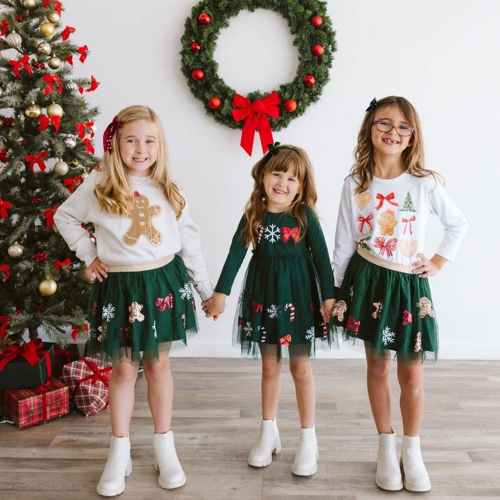 Three children in festive outfits standing in front of a Christmas tree and wreath.