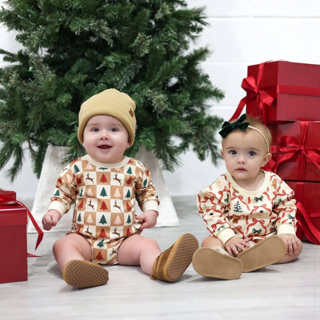 Two babies in Christmas-themed outfits sitting in front of a decorated Christmas tree and presents.