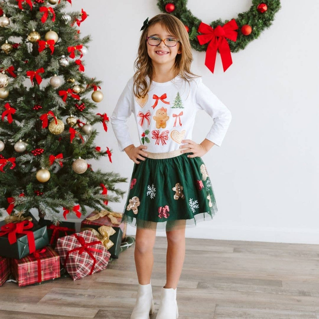 Young girl in festive outfit standing in front of a decorated Christmas tree and wreath.