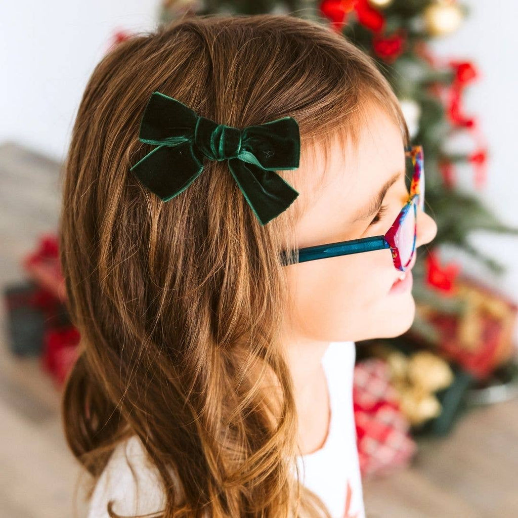 Child with green bow in hair and colorful glasses, Christmas tree in background