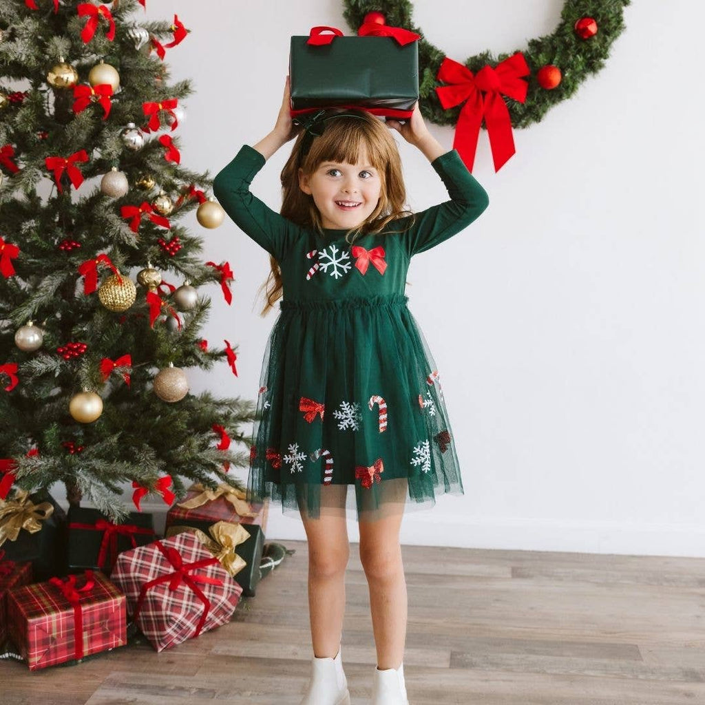 Child in a green Christmas dress holding a gift box, standing in front of a decorated Christmas tree.