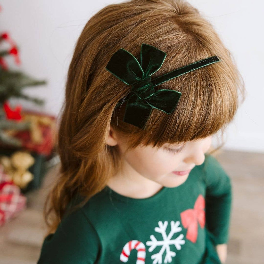Child wearing a green velvet hair bow with a Christmas-themed shirt in a decorated room.