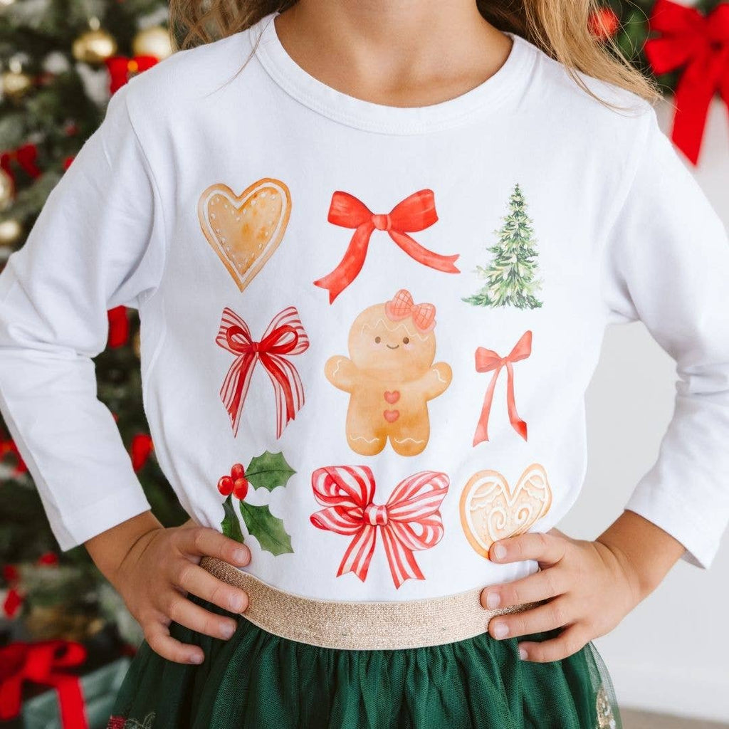 Child wearing a white sweatshirt with Christmas-themed designs in front of a decorated tree.
