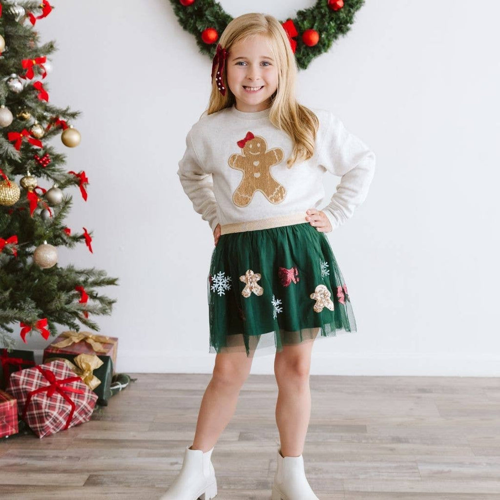 Young girl wearing a Christmas-themed sweater and skirt in front of a decorated Christmas tree.