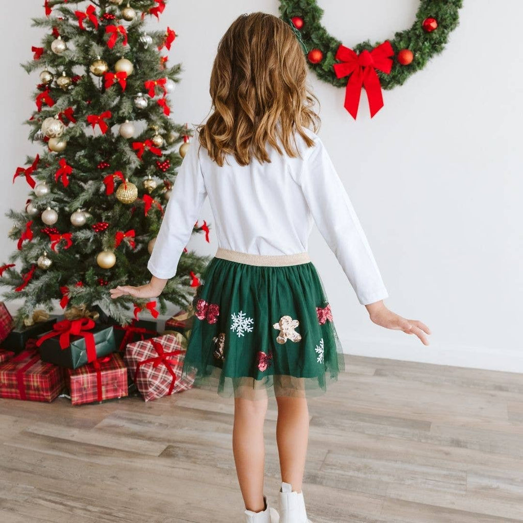 Child in a green skirt with red and white decorations standing in front of a Christmas tree and wreath.