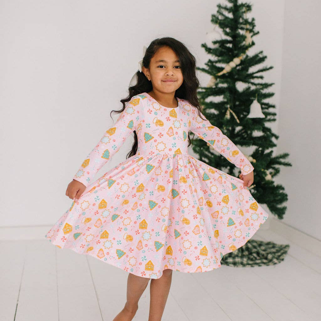 A young girl wearing a long sleeve, full circle twirl skirt dress with a Christmas cookie pattern, standing in front of a decorated Christmas tree.