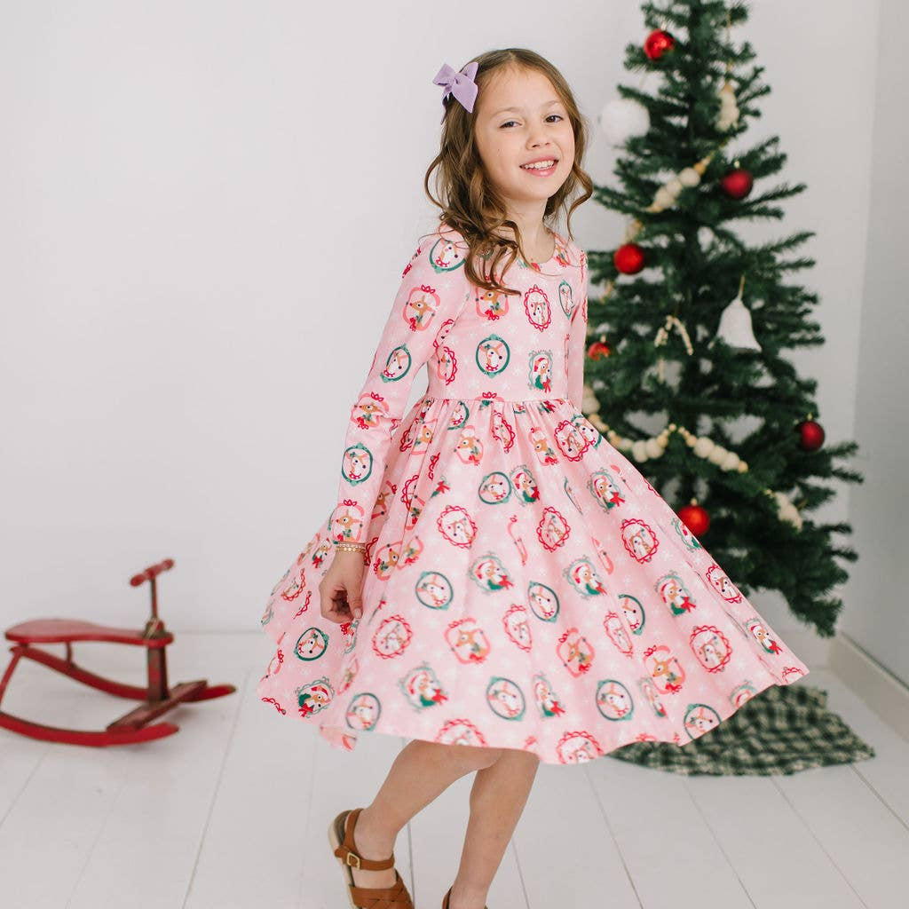 A girl wearing a short-sleeved dress with a reindeer pattern, a scoop back, and a full skirt, standing in a room with a Christmas tree and a red toy cart in the background.