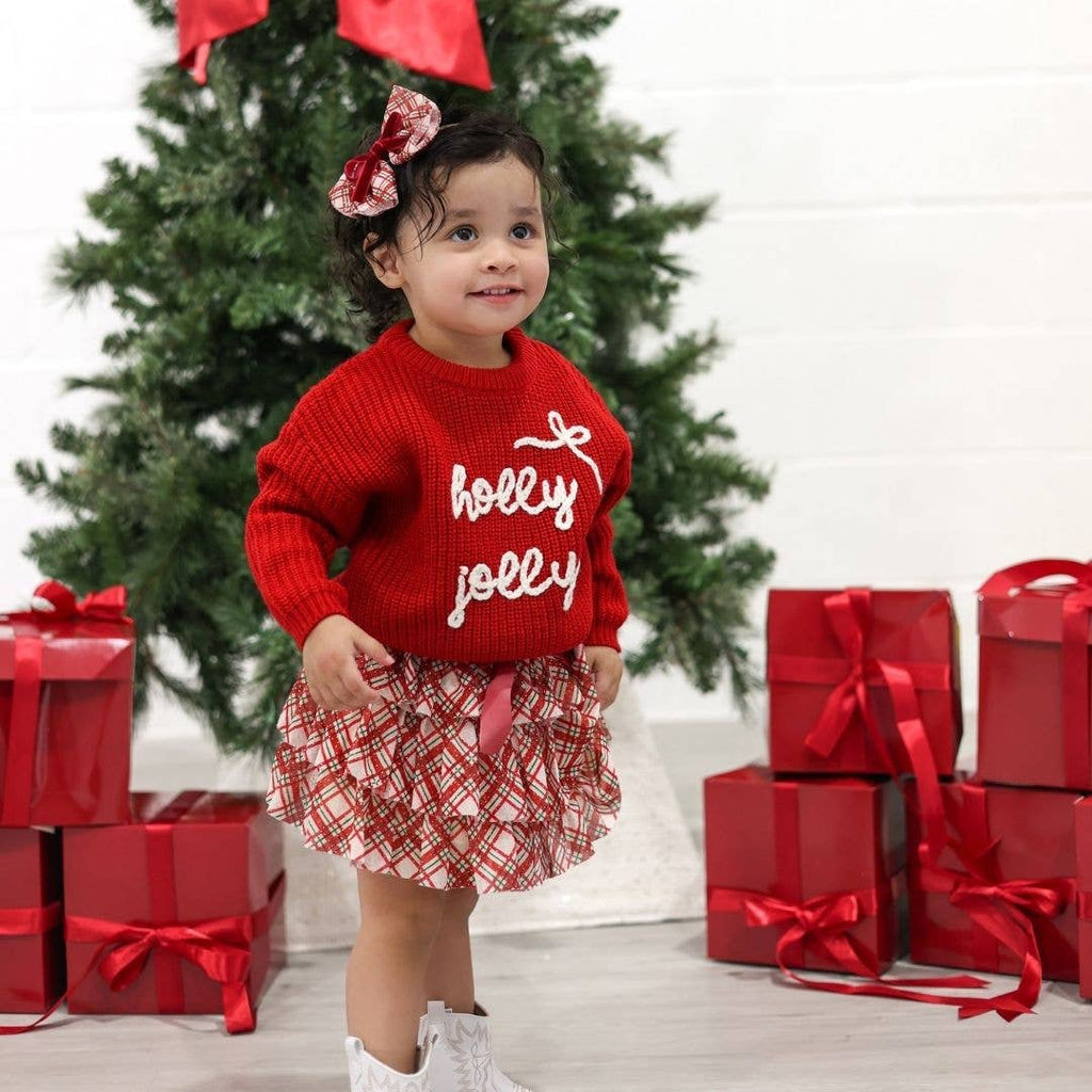 Child wearing a red sweater with 'holly jolly' text, standing in front of a Christmas tree and presents.