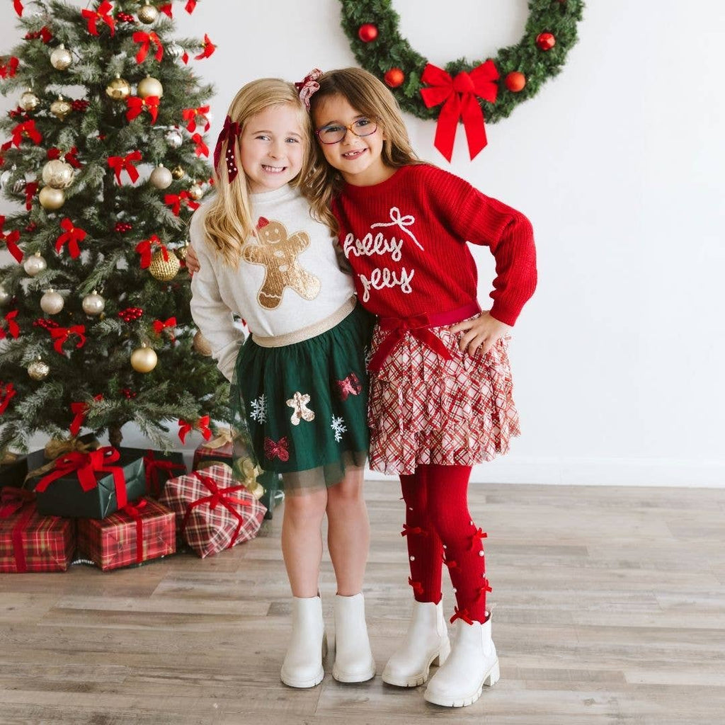 Two children in festive outfits standing in front of a decorated Christmas tree.