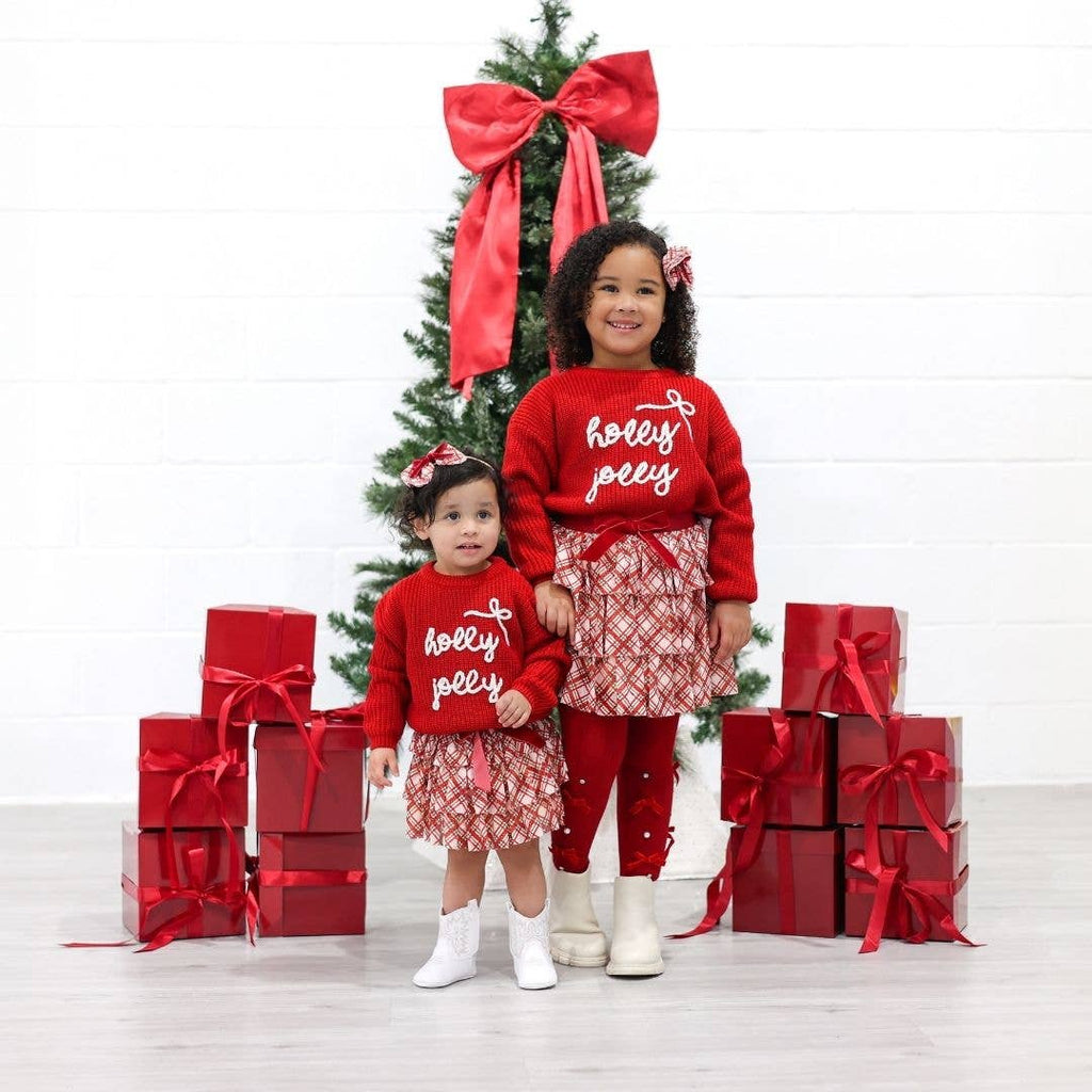 Two children in matching red outfits with 'holly jolly' text, standing in front of a Christmas tree and presents.