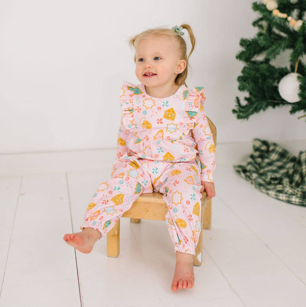 A baby wearing a long-sleeved romper with a Christmas cookie pattern, sitting on a wooden chair, with a decorated Christmas tree in the background.