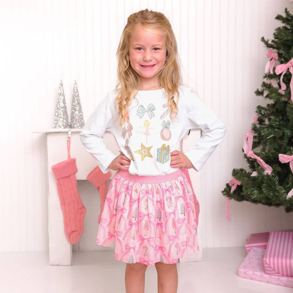 Young girl in a white shirt with colorful designs and a pink skirt standing in front of a Christmas tree and stockings.