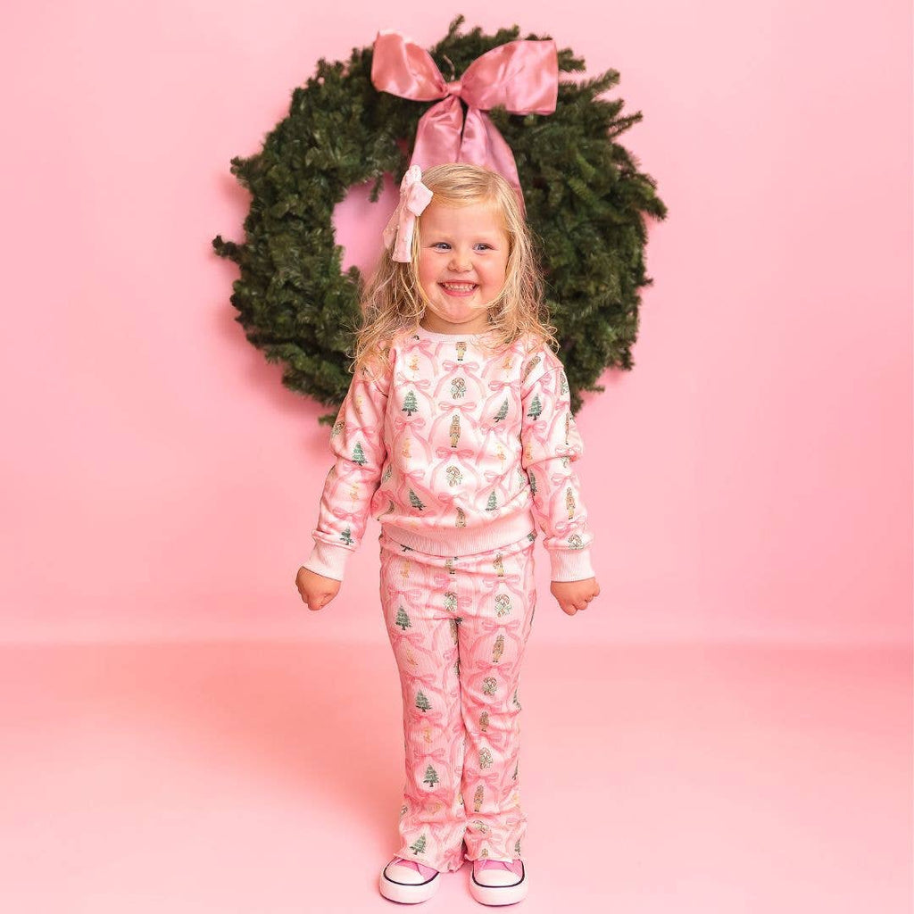 Child wearing pink Christmas-themed pajamas in front of a wreath on a pink background