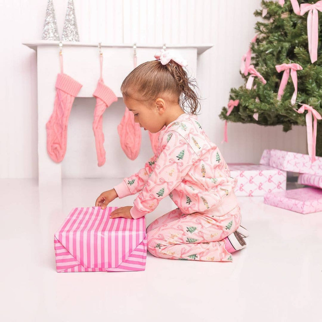 Child in pink pajamas opening a present next to a Christmas tree with pink ribbons.