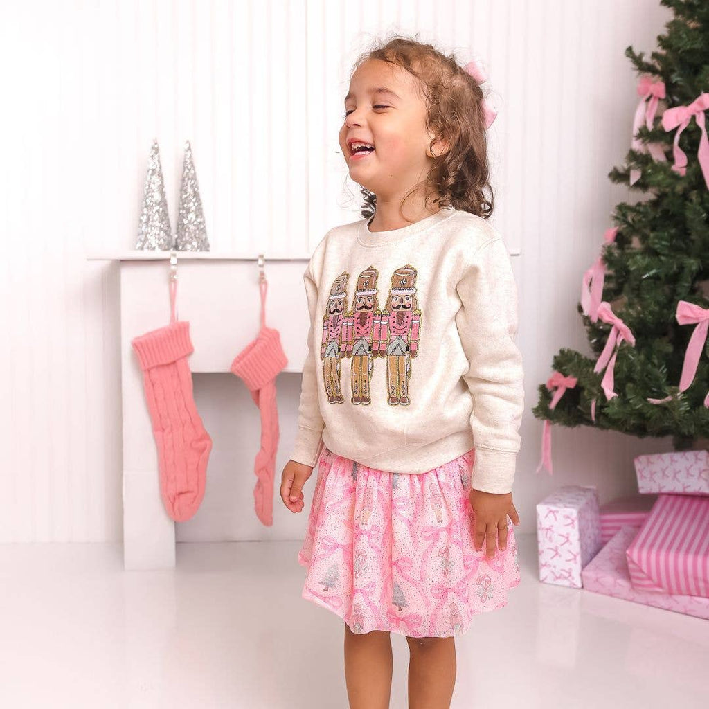Child wearing a white sweater with a Christmas tree design, standing in front of a decorated Christmas tree and stockings.