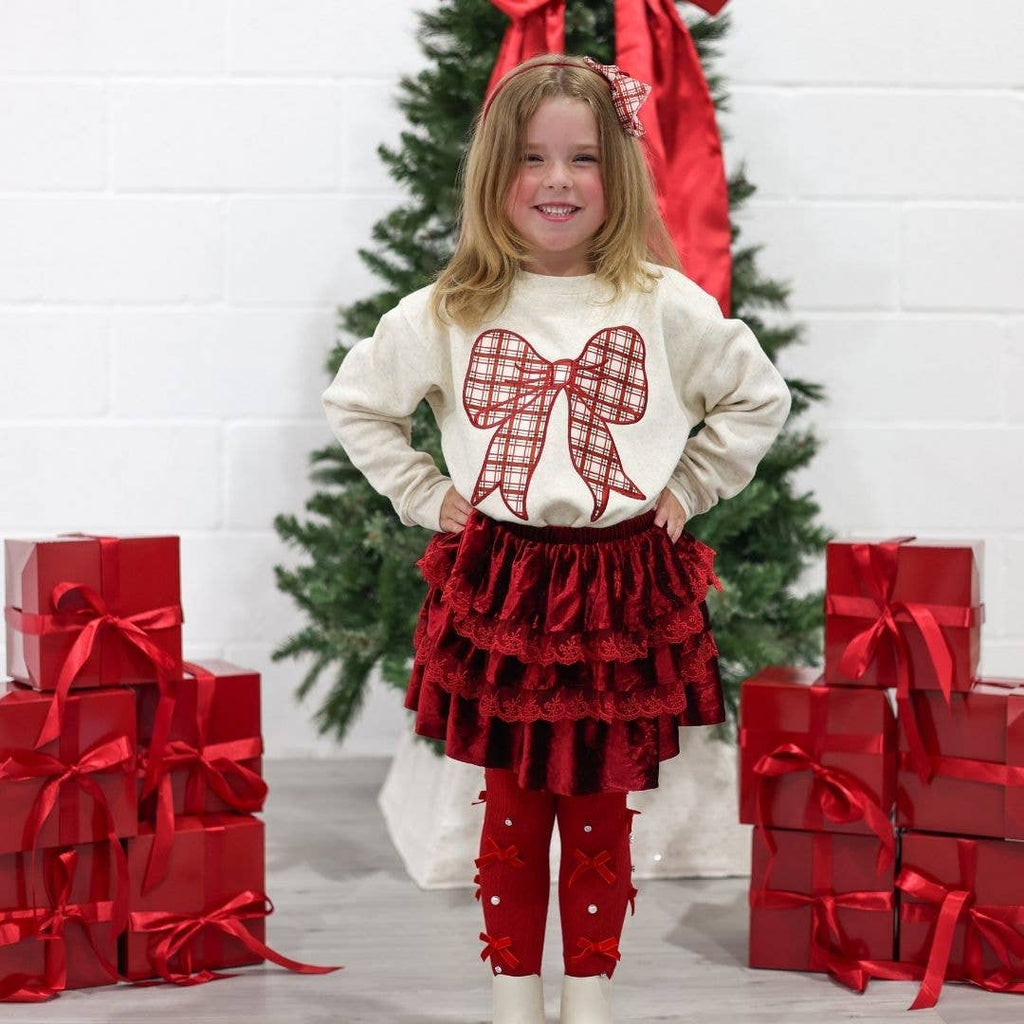 Young girl in festive outfit with red bow shirt and red skirt standing in front of Christmas tree and presents.
