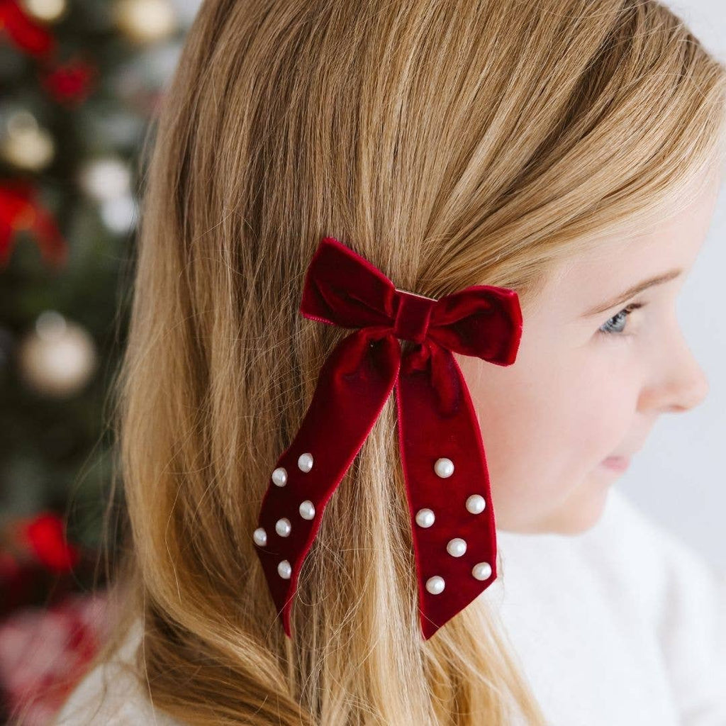 Close-up of a person wearing a red velvet bow with pearl embellishments in their hair, with a blurred festive background.