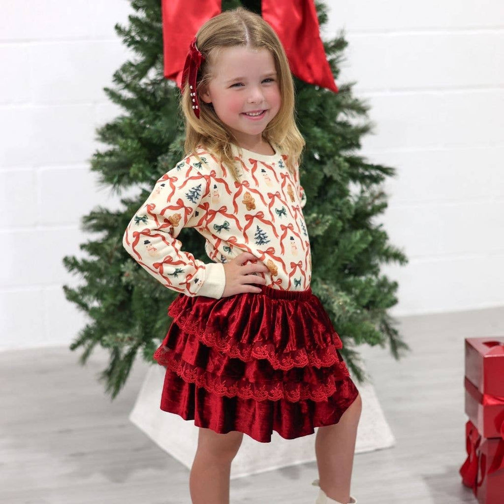 Young girl in a Christmas-themed outfit with a red skirt and patterned top, standing in front of a decorated Christmas tree.
